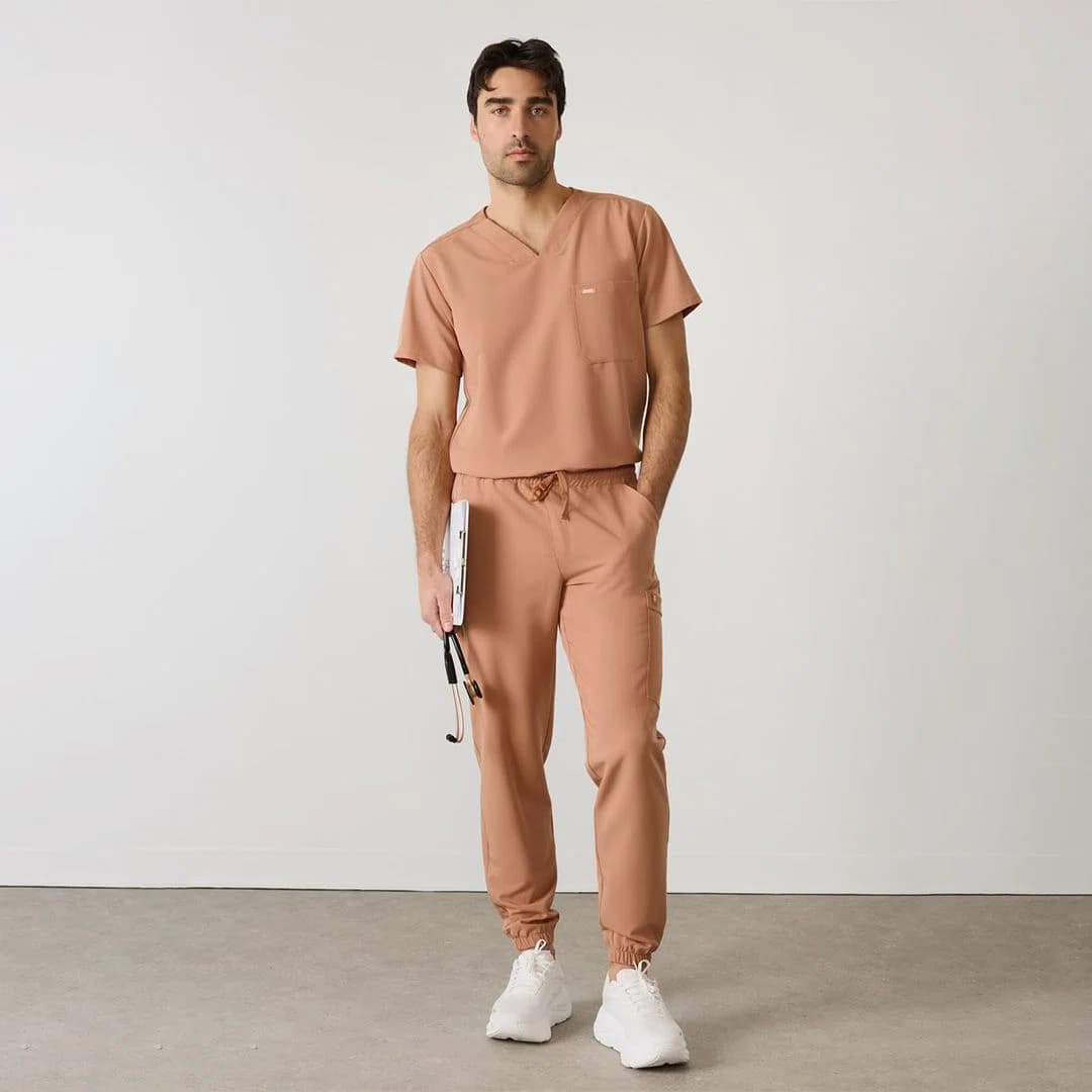 Front view of a modern nurse wearing tailored scrubs with a v-neck top and jogger pants, demonstrating ergonomic movement. The fabric appears breathable and lightweight with a matte finish. An embroidered hospital logo is clearly visible on the chest. The pockets are positioned for easy access to tools. The garment maintains color uniformity under bright clinical lighting.